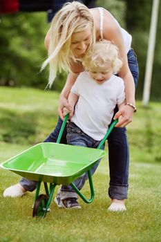 Young mother helping her baby boy in pushing wheelbarrow in garden