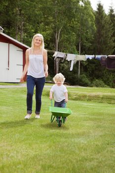 Full length of adorable child pushing a wheelbarrow in garden while walking with mother