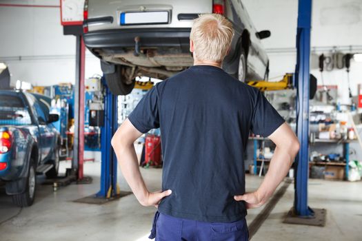 Rear view of car repairer looking at car in auto repair shop with hands on waist