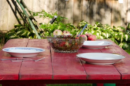 glass dish full of ecologic salad and apples on wooden table in rural house yard.