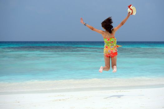 Happy young woman holding hat is jumping on the beach