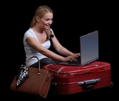 Young blonde woman checking her computer in a hurry on her red suitcase,isolated against a black background.