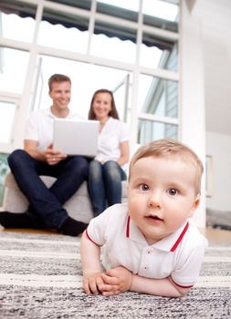 A happy curious young baby boy with parents in the background using computer