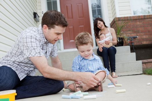 Young Father And Son Drawing With Chalk And Mother Sitting With Baby in Background.