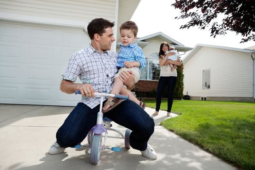 Playful father sitting on tricycle with son while wife standing in background with daughter