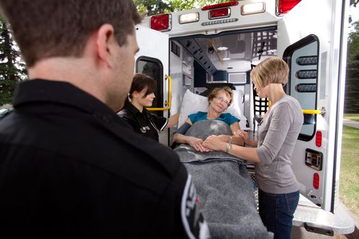 Senior woman being transported to hospital by paramedics, caregiver at side holding her hand