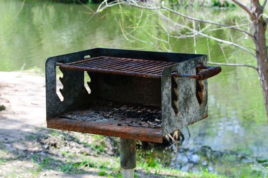 Old rusted charcoal grill near a pond at a park.