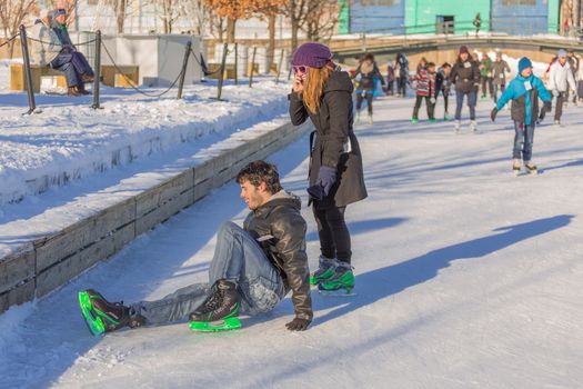  A man has fell and hurt himself while ice skating, and a girl is trying to help him while talking on the phone in a really cold winter day in the Skating Rink in Old Port of Montreal, Quebec ,Canada
