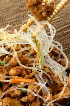 Teriyaki and Picked up Piece of Chicken From Chinese Glass Noodles by Pair of Chopsticks closeup on Straw mat background