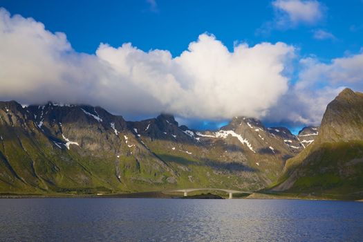 Bridge connecting islands on Norwegian coast with high mountains, patches of snow and peaks in the clouds