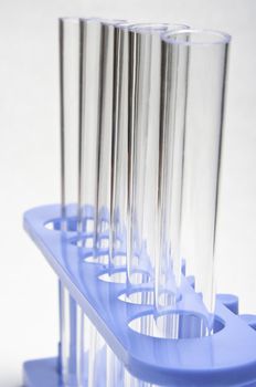 Macro shot of six test tubes, held upright in a blue stand.  Slightly angled and leading away from the viewer.  Soft white background.
