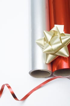 Overhead close up shot of two rolls of Christmas wrapping paper and a gold decorative rosette.  A swirl of shiny red ribbon is placed at the bottom of the frame.  Copy space to left.