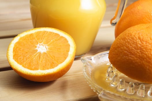 A jug of orange juice with whole and cut oranges and an orange squeezer on a wooden table.