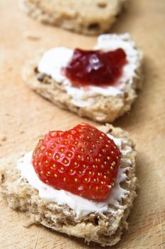 A vertical row of heart shaped granary bread snacks on a wooden chopping board.  Toppings of cream cheese, cut strawberry and jam.