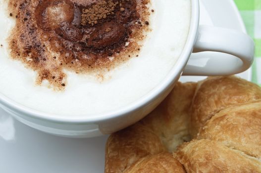 Overhead close up of a cup of coffee with frothed milk and cocoa sprinkles, on a saucer with a croissant.  Gingham tablecloth in background.