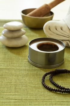 A setting for Buddhist meditation with stacked Zen stones, singing bowl, unlit candle Buddha statue (cropped) and prayer beads on a ribbed green mat.   Light wood table surface just visible at top of frame.