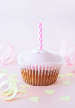 An iced cup cake with birthday candle on pink background with yellow heart confetti and curled paper streamers.