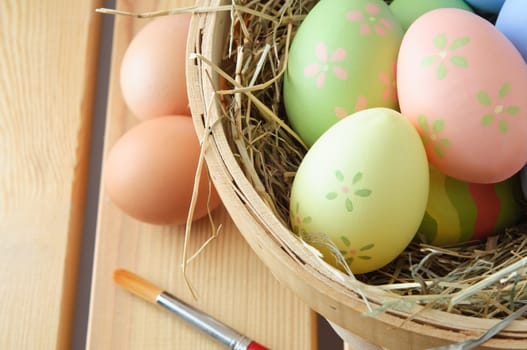 Close up of hand painted Easter eggs in a basket filled with straw with natural, unpainted eggs and paintbrush resting on a wood plank table top.