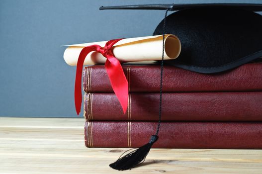 Graduation mortarboard and scroll tied with red ribbon on top of a stack of old, worn books on a light wood table.  Grey background.  