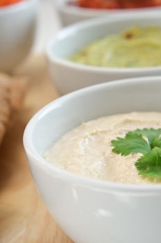 Close up of a bowl of hummus with guacamole and salsa dips in the background.  Pitta bread just visible at left frame.