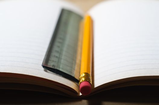 An opened student's exercise book on a wooden table, blank pages revealed.  A pencil and ruler lie vertically in the centre seam. Horizontal (landscape) orientation.