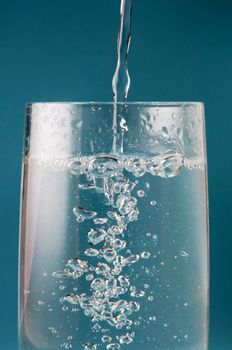 Vertical action shot of a stream of water pouring into a tall glass, creating oxygen bubbles.  Dark turquoise background.