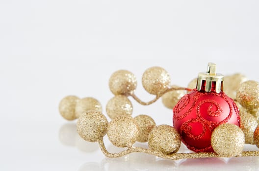 A red Christmas bauble with glittery spiral pattern and gold glitter ball decorations on an off-white reflective surface with copy space above and to left.