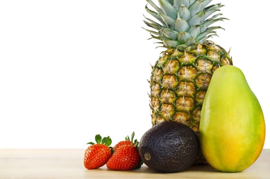 A fruit composition  of pinapple, mango, avocado and strawberries on a light wooden table against a white background.  Copy space to left.