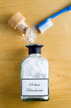 A toothbrush and a  bottle of Sodium Bicarbonate (baking soda) laid flat and shot from overhead on a wooden table.  Cork open and powder spilling on to table.  