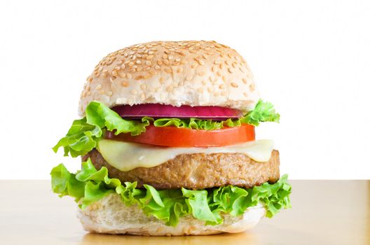 A veggie burger (made from soya protein) in a sesame seed bap with layers of curly lettuce, melted cheese, tomato and onion, on a light wood table with white background.