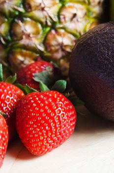 Fruit composition on light wood with group of ripe, red strawberries and avocado to the fore, and a pineapple in soft focus background.