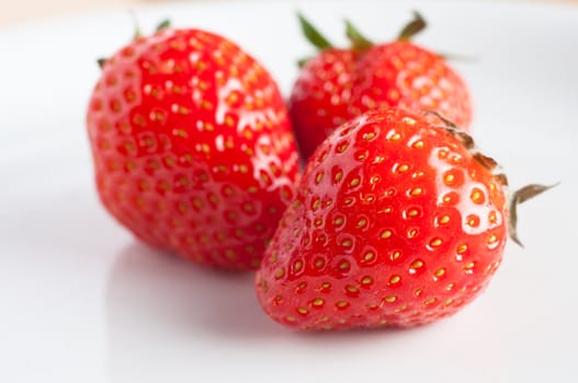 Close up of a group of three fresh strawberries on a white plate, reflection visible.