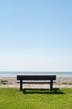 Rear view of an empty bench facing the sea.  Grass behind and shingle beach in front, leading to sea.  Upper two thirds provides ample copy space bright blue sky.