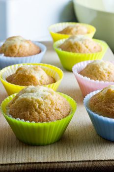 Rows of cupcakes, fresh from the oven, in colourful silicone baking cups on a wooden board with mixing bowl in the background.