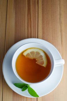 Overhead shot of lemon tea in a white cup and saucer with half a lemon slice and mint leaves on a wooden planked table.