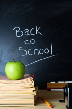 Teacher's desk with a pile of books, an apple and other equipment.  The words 'Back to School' written in chalk on the blackboard in soft focus background.