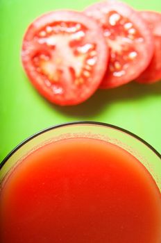 Overhead close up of a glass of tomato juice with tomato slices in soft focus background on green surface.  