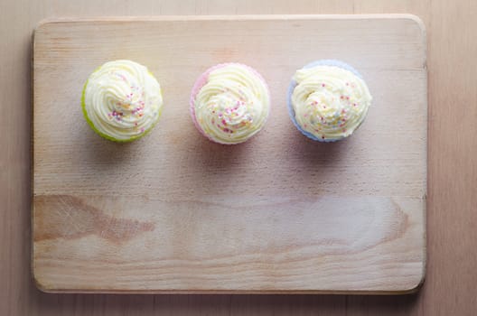 Overhead shot of three cupcakes on a wooden chopping board, topped with swirls of buttercream icing and colourful sprinkles.