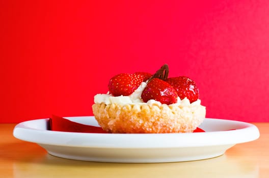 A strawberry and cream pastry cake with chocolate decoration on red napkin and white plate, against a red background.