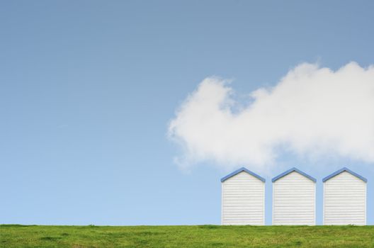 A row of three beach huts on a grassy hill against a bright blue sky with a big white fluffy cloud.  Copy space above and left.