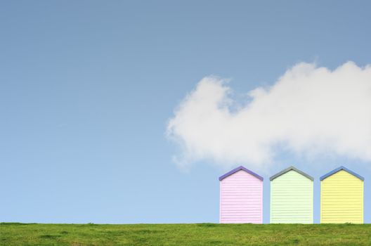 A row of three colourful beach huts on top of a grassy hill against a bright blue sky with white fluffy cloud. Copy space above and left.
