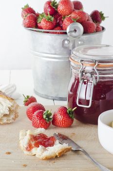 A farmhouse kitchen table scene of freshly made strawberry jam in storage jar with a tin pail of strawberries in the background and bread and strawberries on a wooden chopping board.