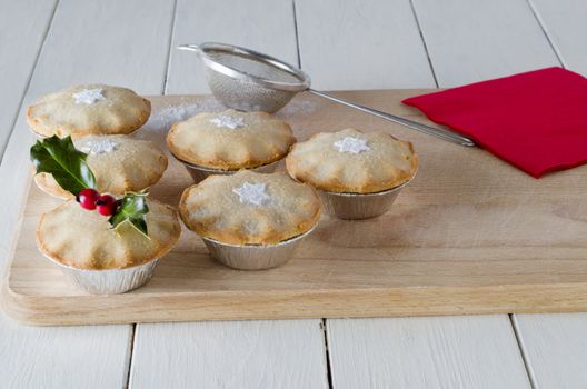 Mince pies on a wooden board, decorated with star shapes and holly.  A red napkin, old tea strainer (sieve) and scattered icing sugar rest on the board which sits on a white, planked table.