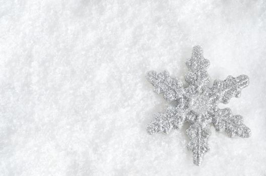 Winter background shot from above.  Glittery silver Christmas snowflake faces upwards on lower right, lying in fake snow.