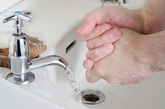 Male hands being washed in old china sink with water running from stainless steel tap, and a bar of soap resting on a wooden soap dish.