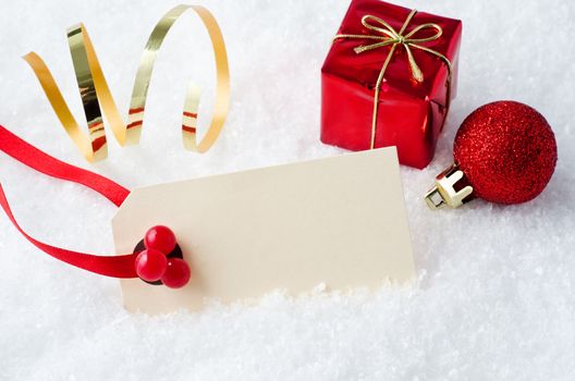 A blank cream coloured Christmas tag on red ribbon with artificial holly berries, partly submerged in fake snow with a gold foil twist, a gift box and a sparkly bauble behind.