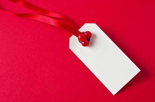 A blank Christmas gift tag label, attached to a shiny red ribbon with artificial red holly berries, on a red background with copy space.