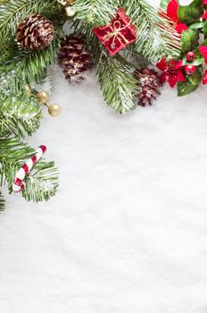 A Christmas border to left and top of frame consisting of artificial foliage, real pine cones and decorative ornments, sprinkled with snow on a fake snow background.   Snow provides copy space.