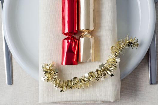 Overhead shot representing Christmas dinner, with red and gold crackers and tinsel laid on a napkin, with plate, cutlery and tablecloth underneath.