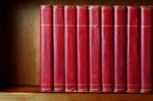 A row of old, battered, matching encyclopaedias (circa 1950s) lined up on a shelf, with titles removed to leave blank spines.  Red leather effect with gold striped trims.  Shelf has been darkened artificially to give impression of age.